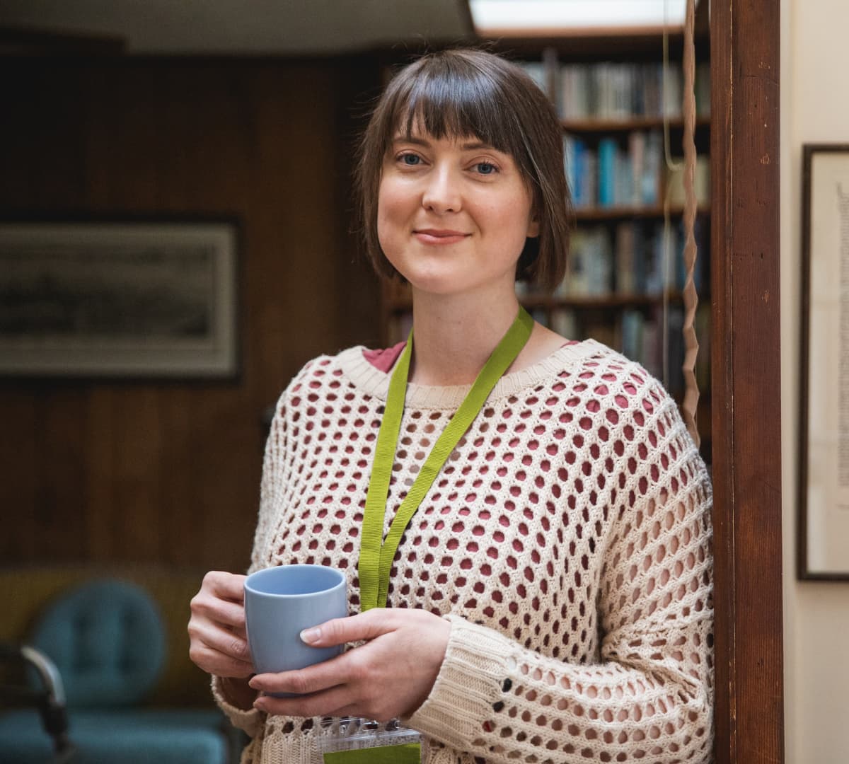 A woman with black hair and wearing a green lanyard standing inside the house while holding a cup of coffee and smiling