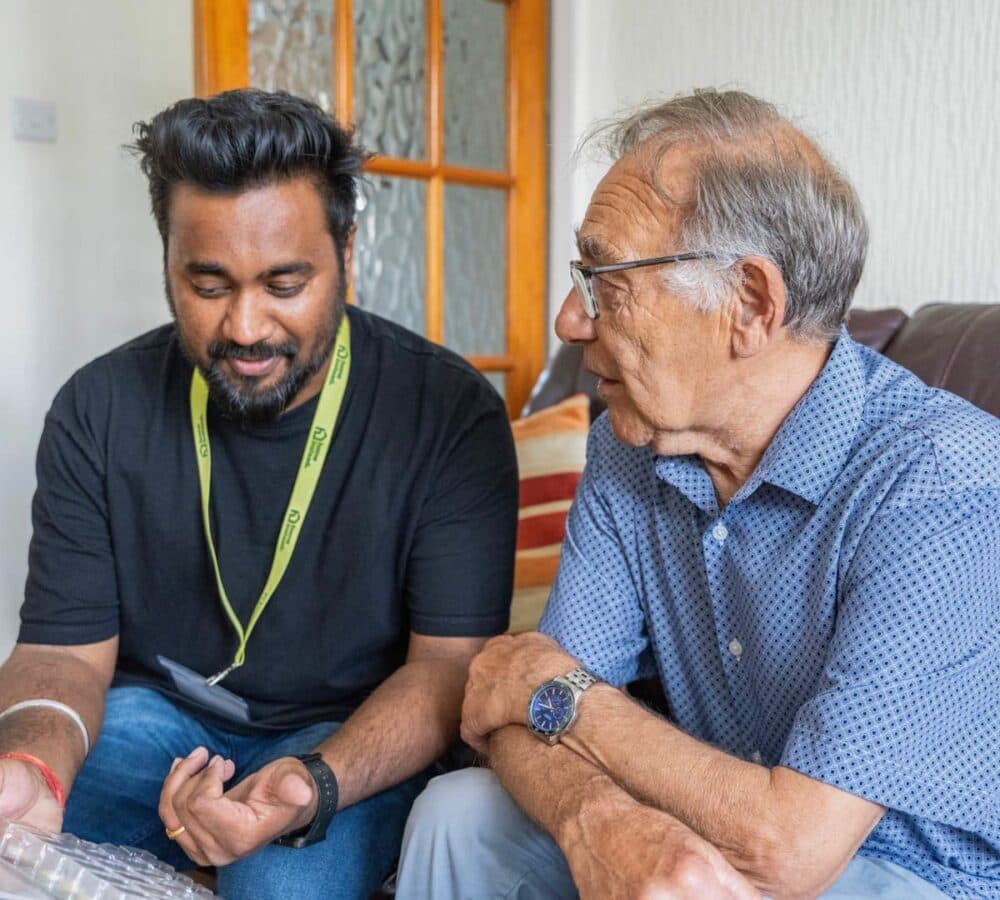 An older male adult with grey hair chatting with his younger male carer inside the house