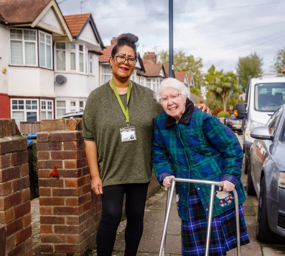 An older female adult with white hair happy and smiling while walking and using a walker with her younger female carer with black hair and wearing eyeglasses outdoors with houses at the background