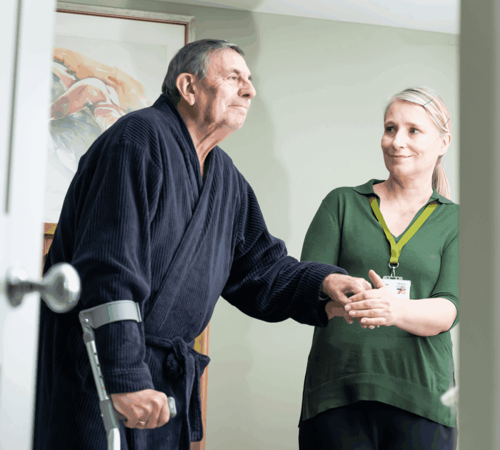 Older man with crutch receives help from a smiling caregiver in a green top inside a room. - Home Instead