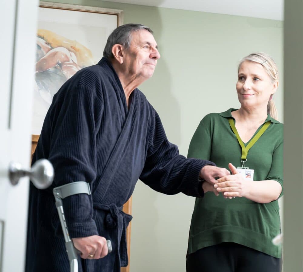 An older male adult with grey hair and wearing a robe walking using a crutch while being helped by a female carer wearing green