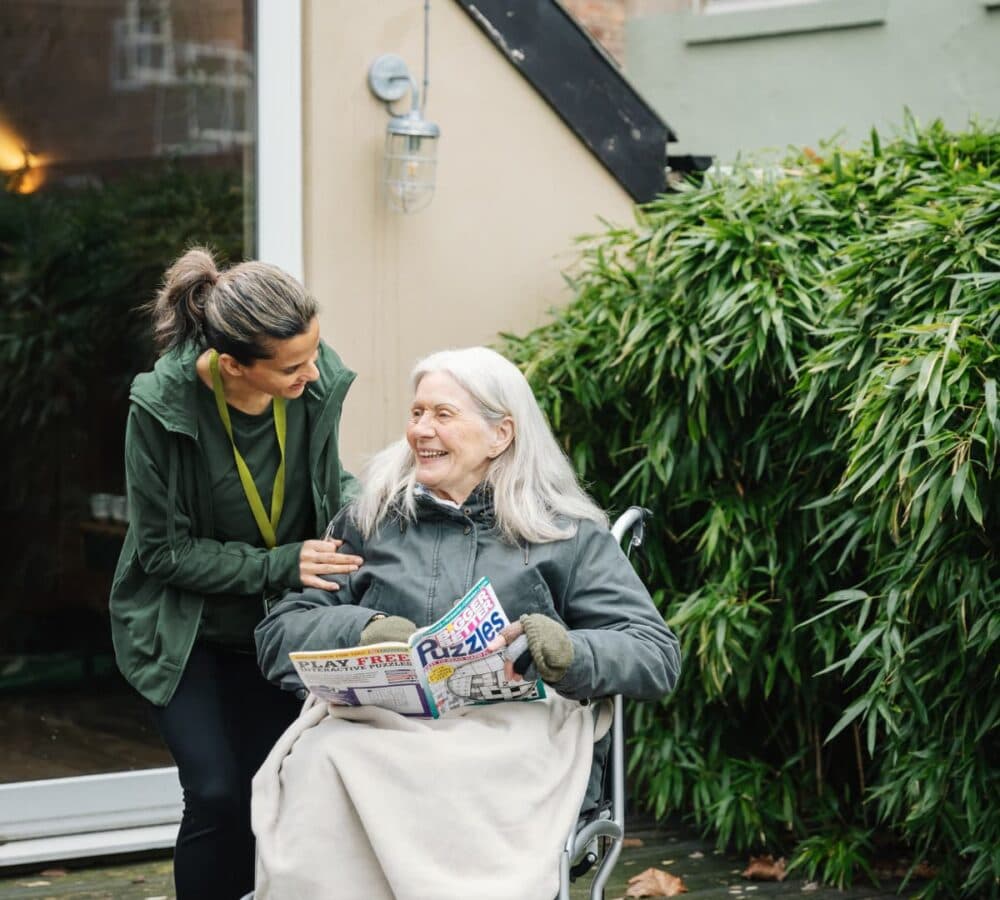 An older female adult with white long hair outdoors using a wheelchair with her younger female carer wearing green