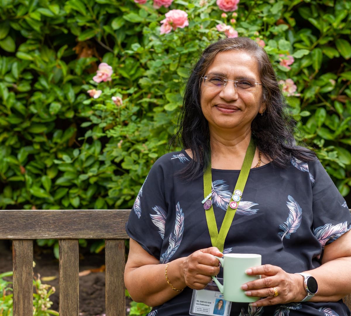A woman with long black hair and wearing eyeglasses happy and smiling while holding a cup of coffee while sitting on the bench inside the garden
