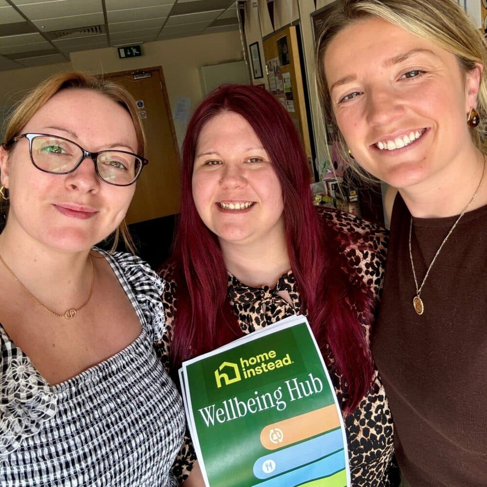 Three women smiling indoors, one holding a "Home Instead Wellbeing Hub" sign. - Home Instead