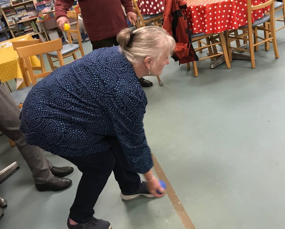 An elderly woman bends down indoors, holding a small blue ball, with tables and chairs in the background. - Home Instead