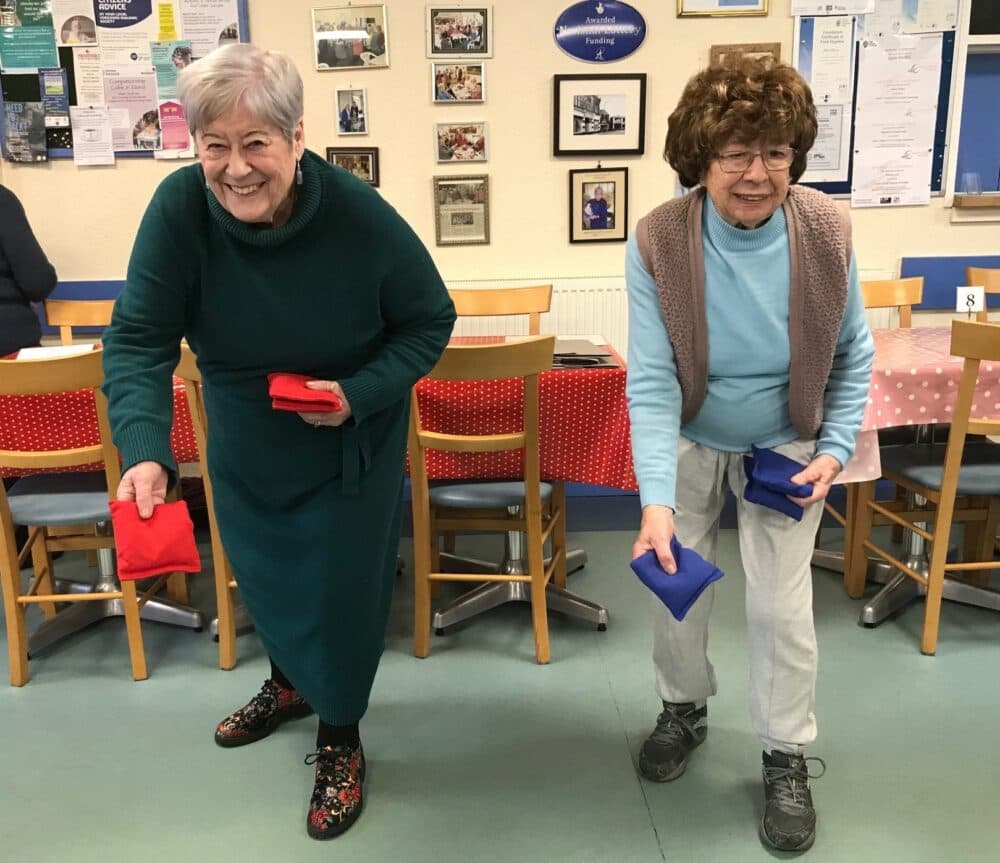 Two older women smiling and playing a bean bag toss game indoors at a community center. - Home Instead