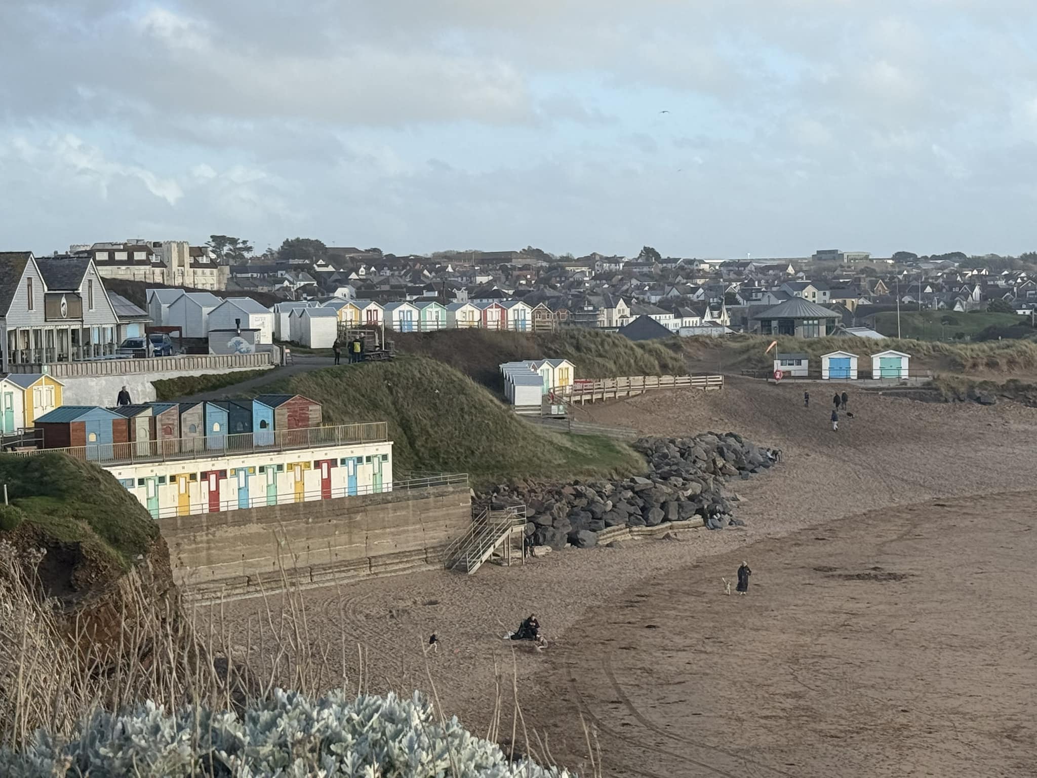 Beach huts and houses overlook a sandy beach with a few people walking along the shore. - Home Instead