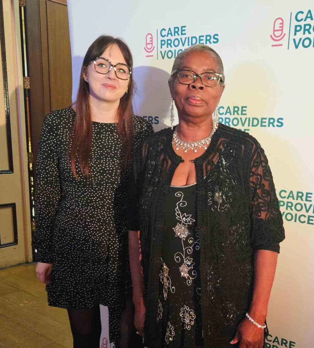 Two women stand in front of a "Care Providers Voice" backdrop, dressed in formal black attire and smiling. - Home Instead