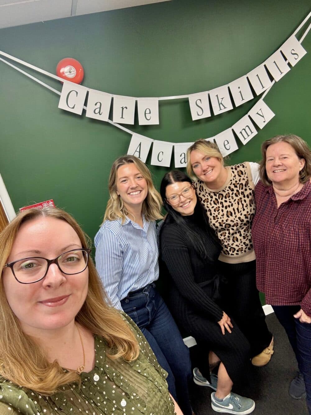Five women smiling in front of a green wall with a "Care Skills Academy" banner hanging behind them. - Home Instead