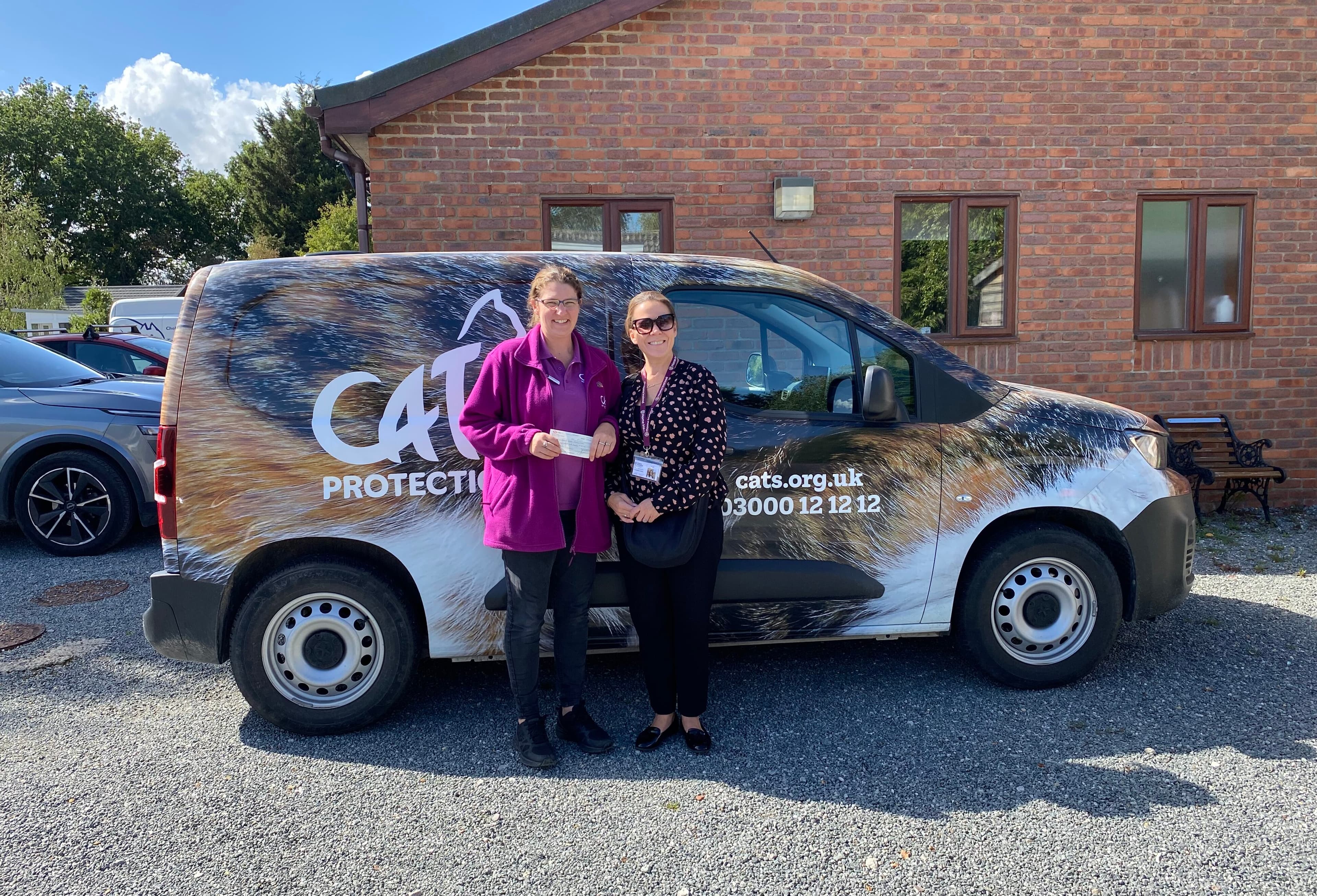Two women stand smiling in front of a Cats Protection van parked beside a brick building on a sunny day. - Home Instead