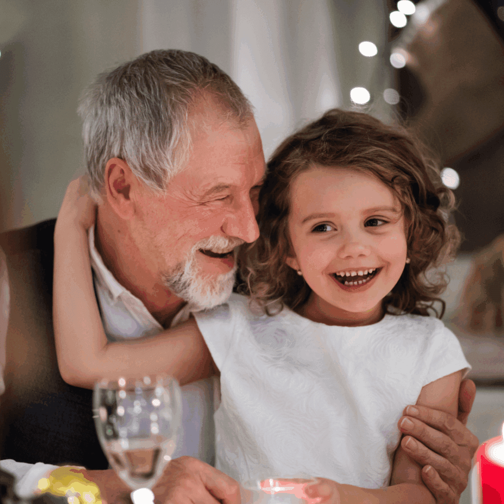 Smiling older man hugs a laughing young girl at a festive, warmly lit dinner table. - Home Instead