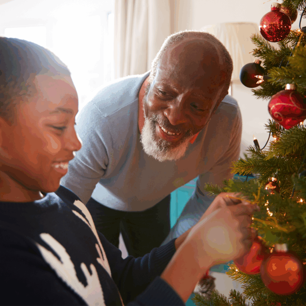 Smiling grandfather and grandson decorate a Christmas tree together with red ornaments in a cozy living room. - Home Instead