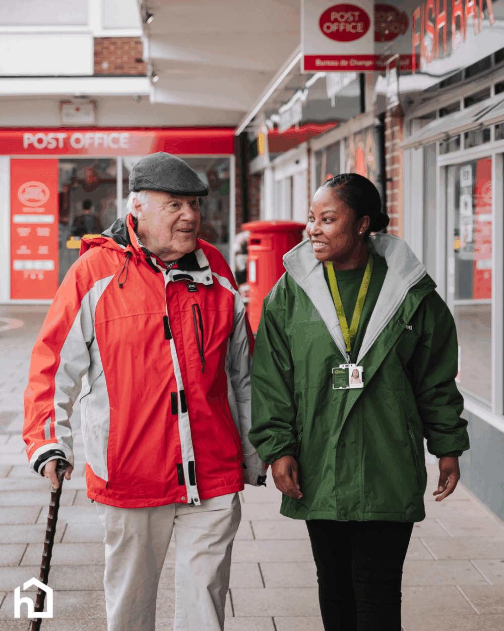 A care worker walking and talking with an older man outside a post office, both smiling warmly.