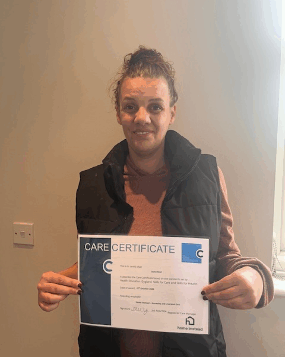 A woman smiling and holding a care certificate while standing indoors near a light-colored wall. - Home Instead