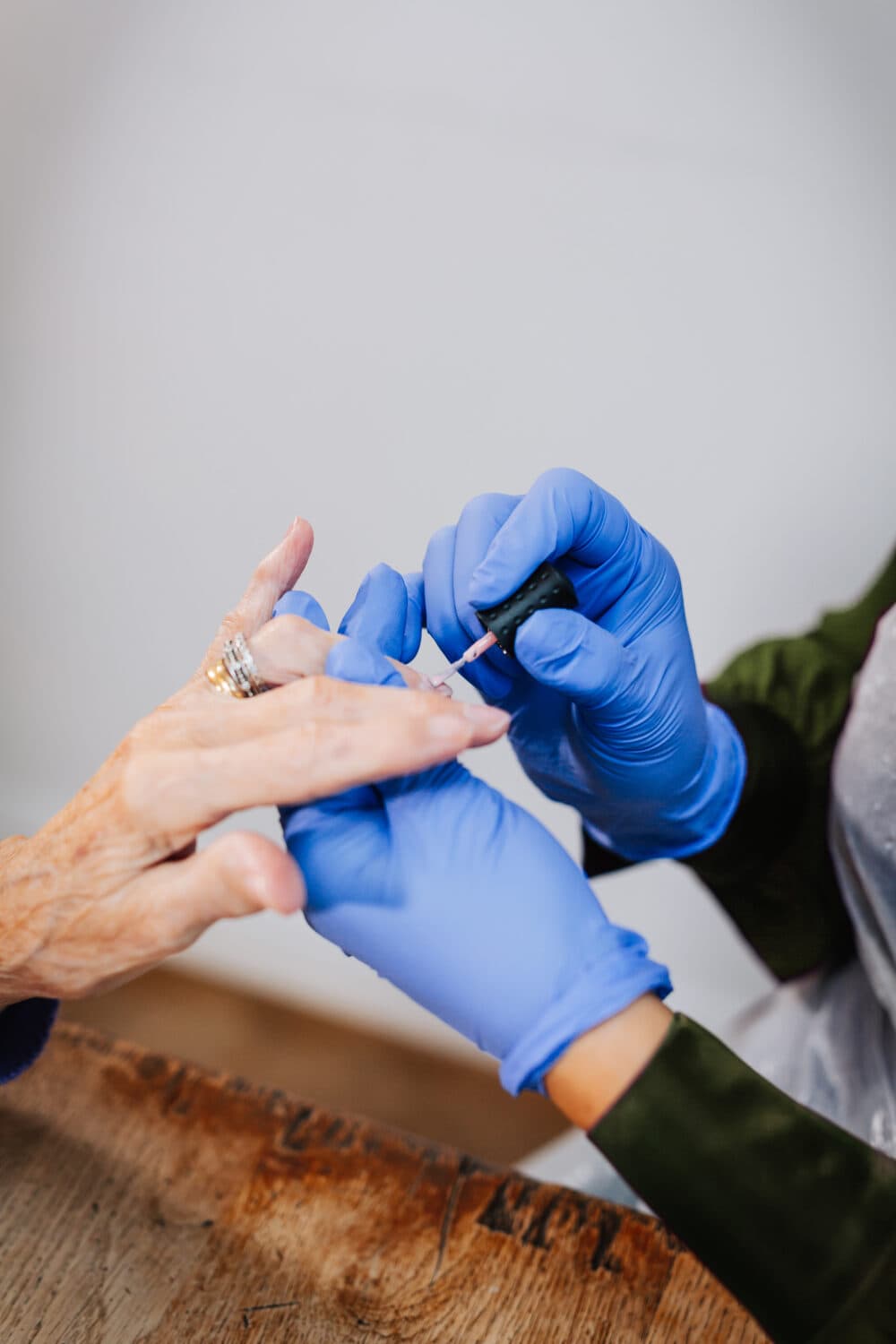 A person wearing blue gloves applies nail polish to another person's fingernails at a wooden table. - Home Instead