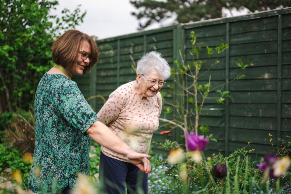 Two women, one elderly, smiling and looking at flowers together in a garden with a green wooden fence behind them. - Home Instead