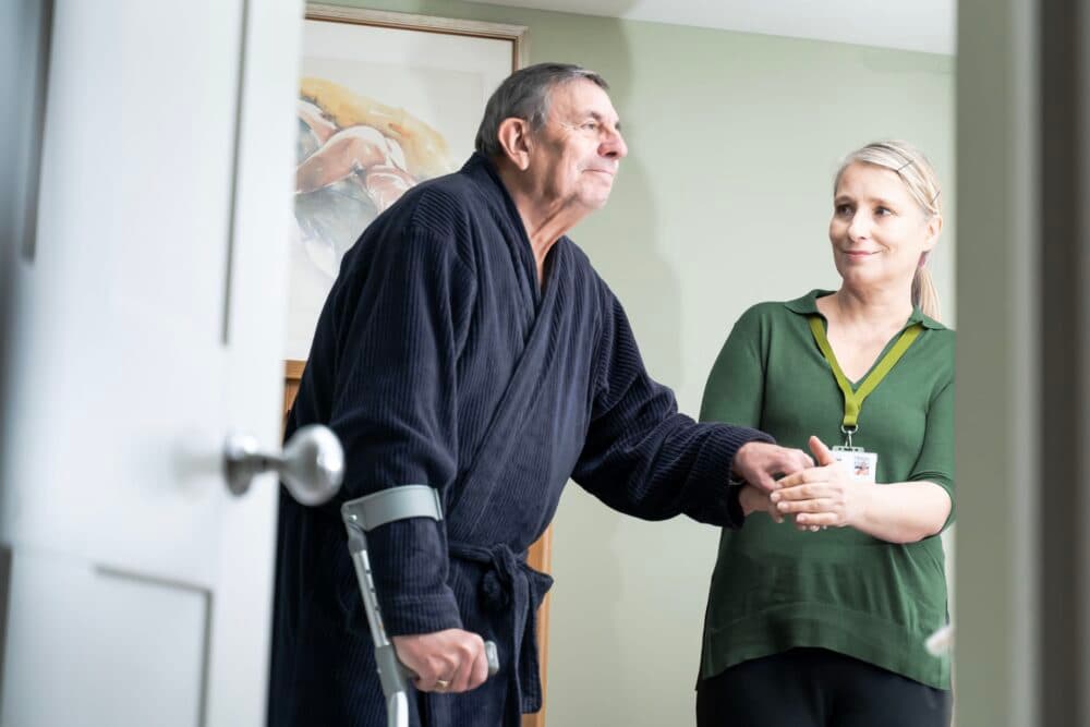 An elderly man with crutches is helped by a caregiver in a green shirt inside a bright room. - Home Instead