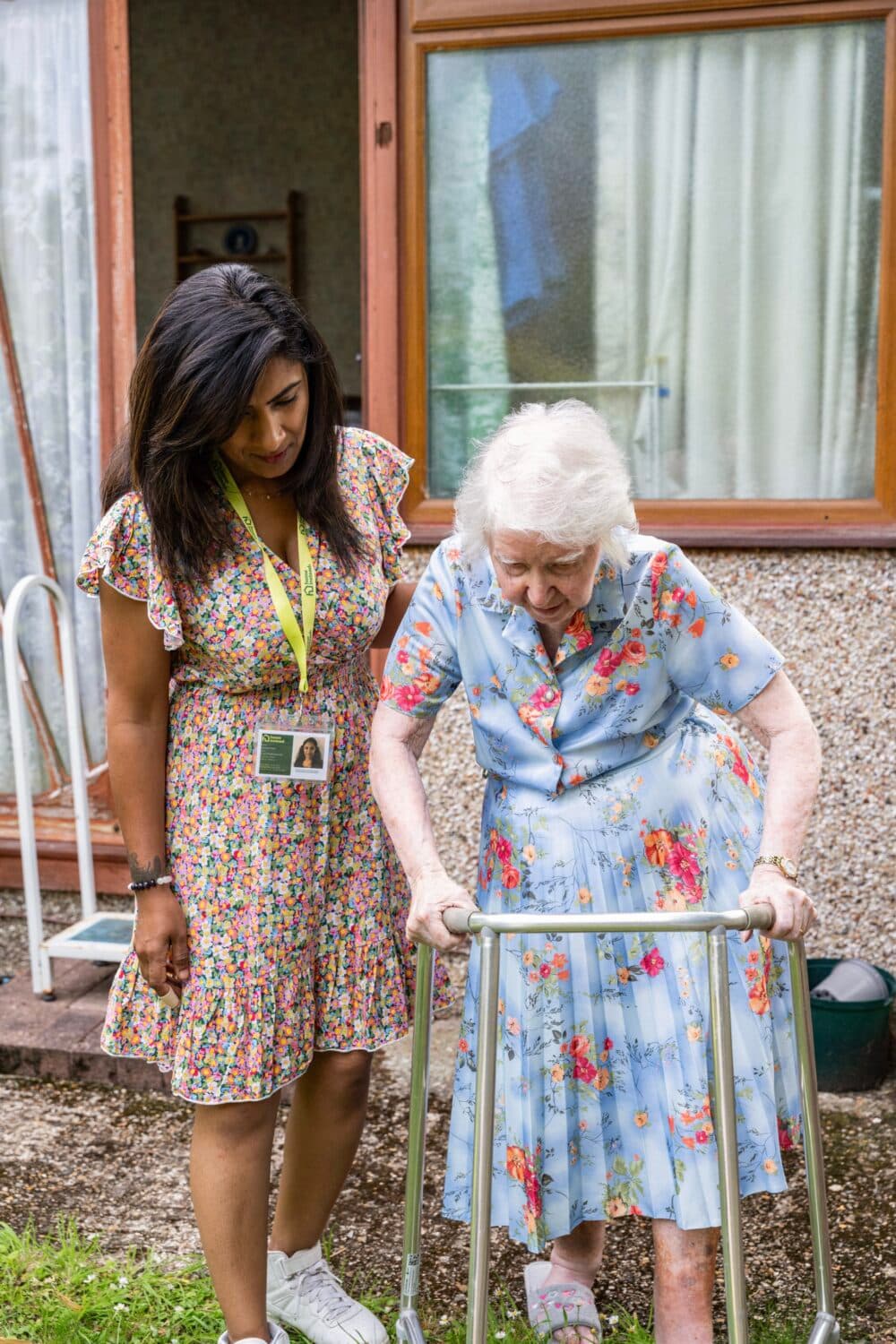 A young caregiver supports an elderly woman with a walker outside a house, both wearing floral dresses. - Home Instead
