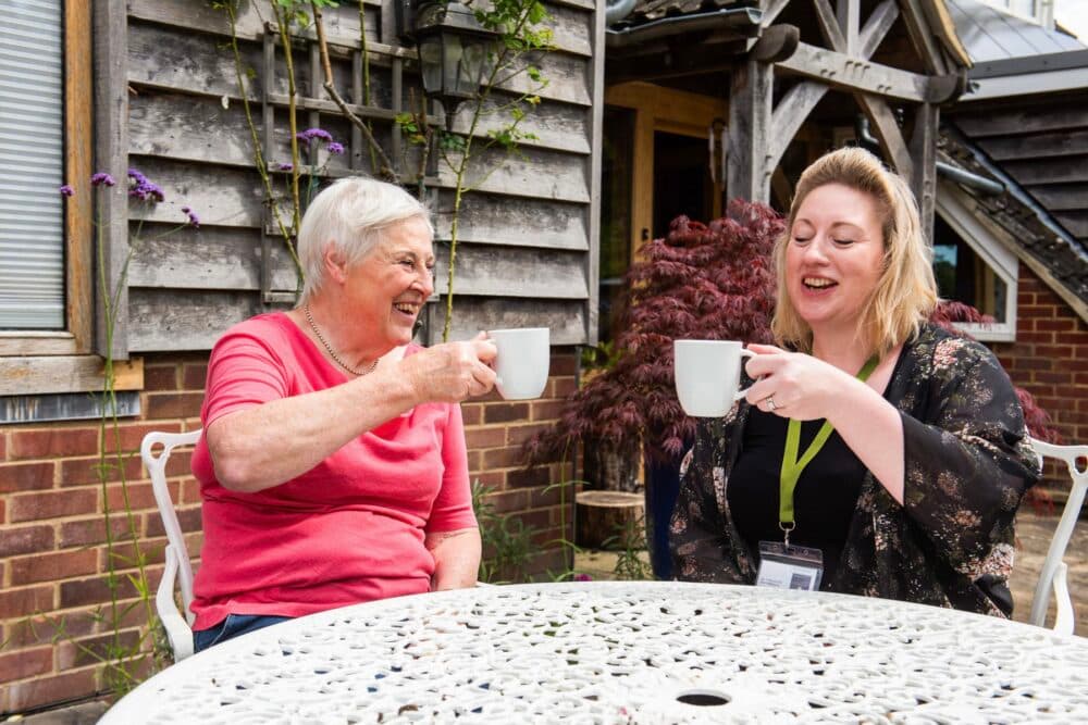 Two women sit outside at a table, smiling and holding mugs, enjoying a drink together. - Home Instead