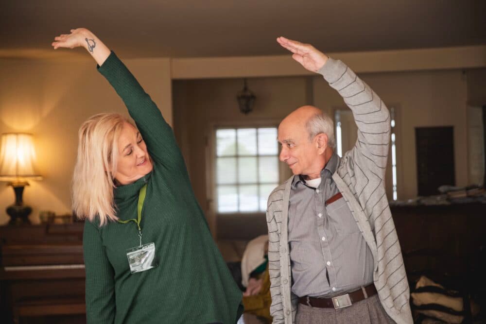 An elderly man and woman do side stretches together in a cozy, warmly lit living room. - Home Instead