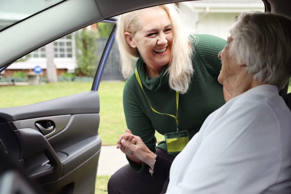Woman helps elderly lady in car, both smiling warmly, suggesting care and support in a residential area. - Home Instead