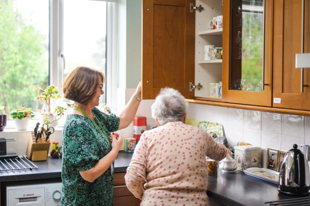 Two women in a kitchen, one opening a cupboard, the other reaching for something on the counter. - Home Instead