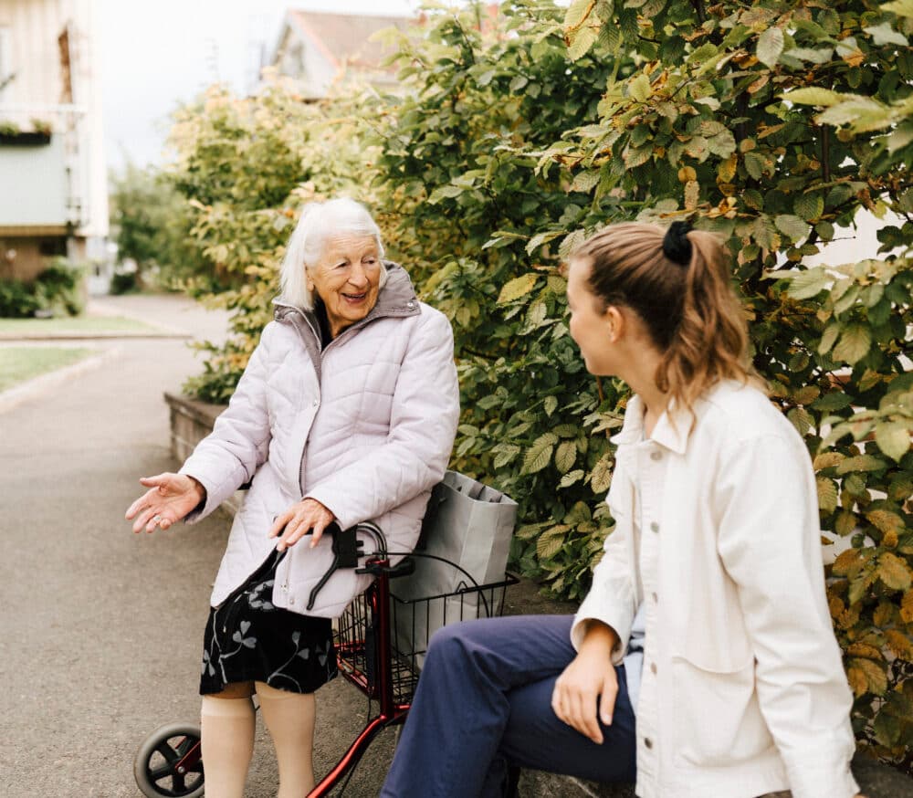 An elderly woman with a walker talks to a younger woman while sitting outside near bushes. - Home Instead