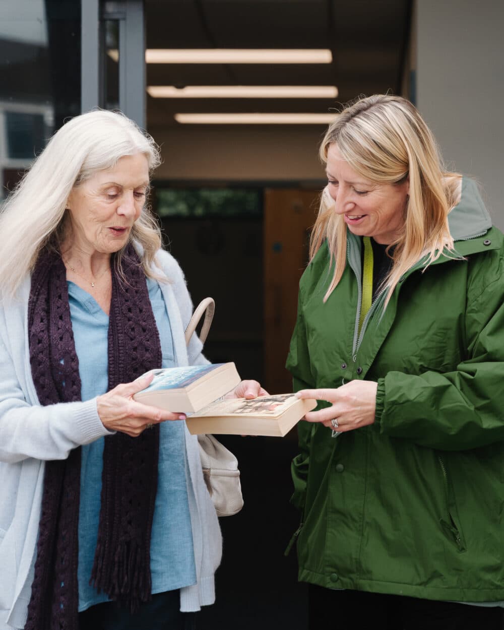 Two women standing outside, smiling and exchanging books with each other. - Home Instead