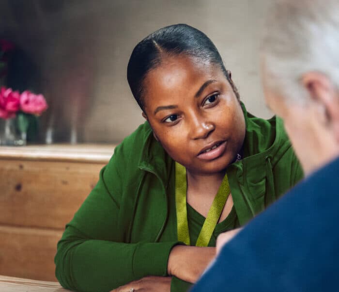 A woman in a green jacket listens attentively to an older person at a wooden table indoors. - Home Instead