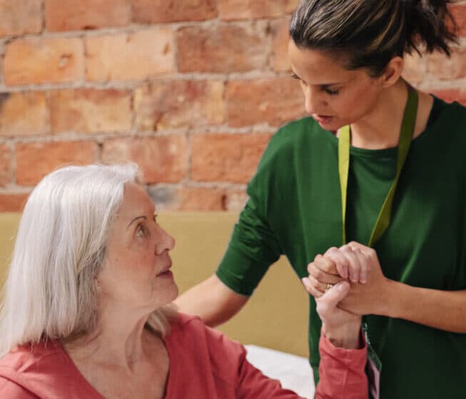 A caregiver in green comforts and holds hands with an elderly woman with gray hair sitting indoors. - Home Instead