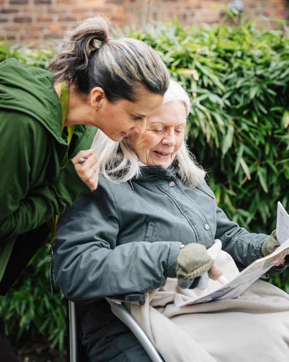 A young woman and an older woman read a newspaper together outdoors, smiling and bundled in jackets. - Home Instead