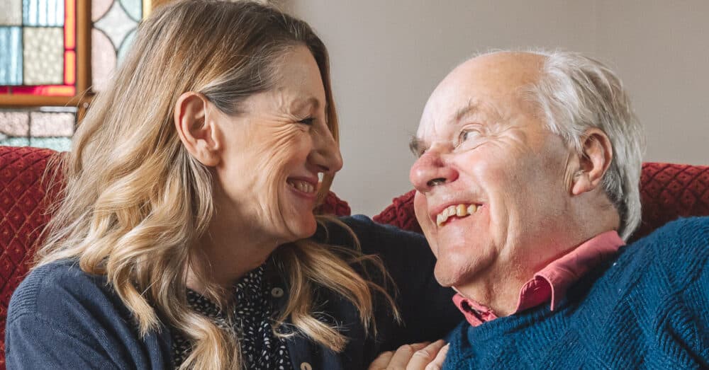 A smiling woman and older man sit close together, sharing a joyful moment indoors. - Home Instead