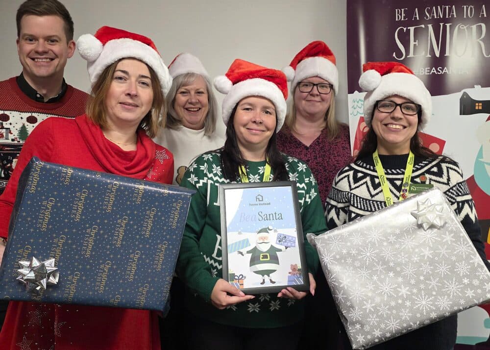 Six people wearing festive sweaters and Santa hats holding wrapped gifts and a "Be a Santa" sign. - Home Instead
