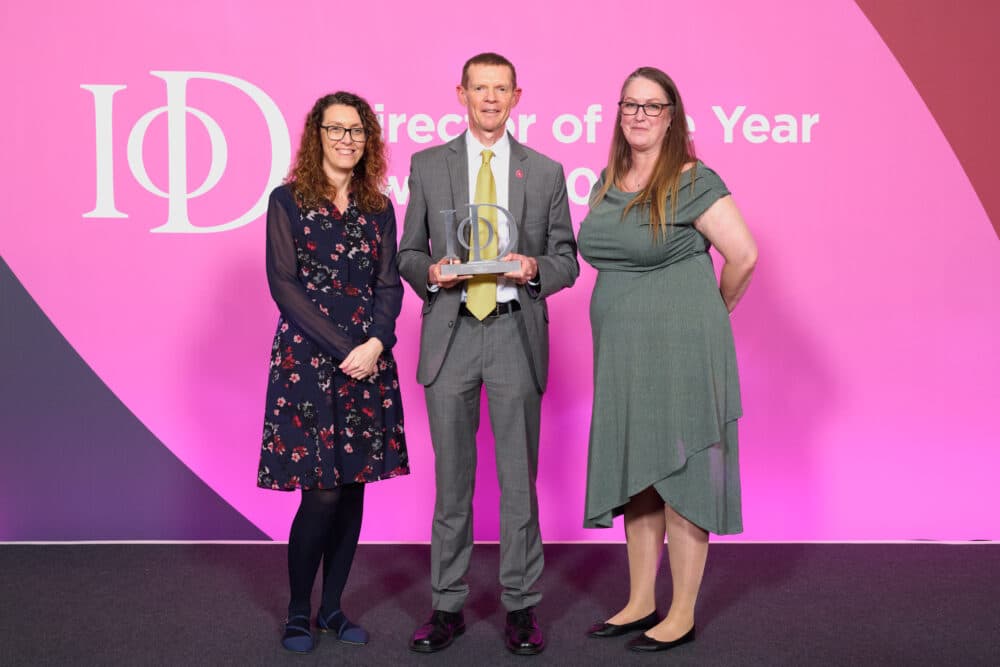 Three people stand in front of a pink backdrop, with the man in the center holding an award. - Home Instead