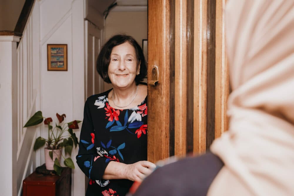 Smiling woman in a floral top opens her front door to greet a guest wearing a beige headscarf. - Home Instead