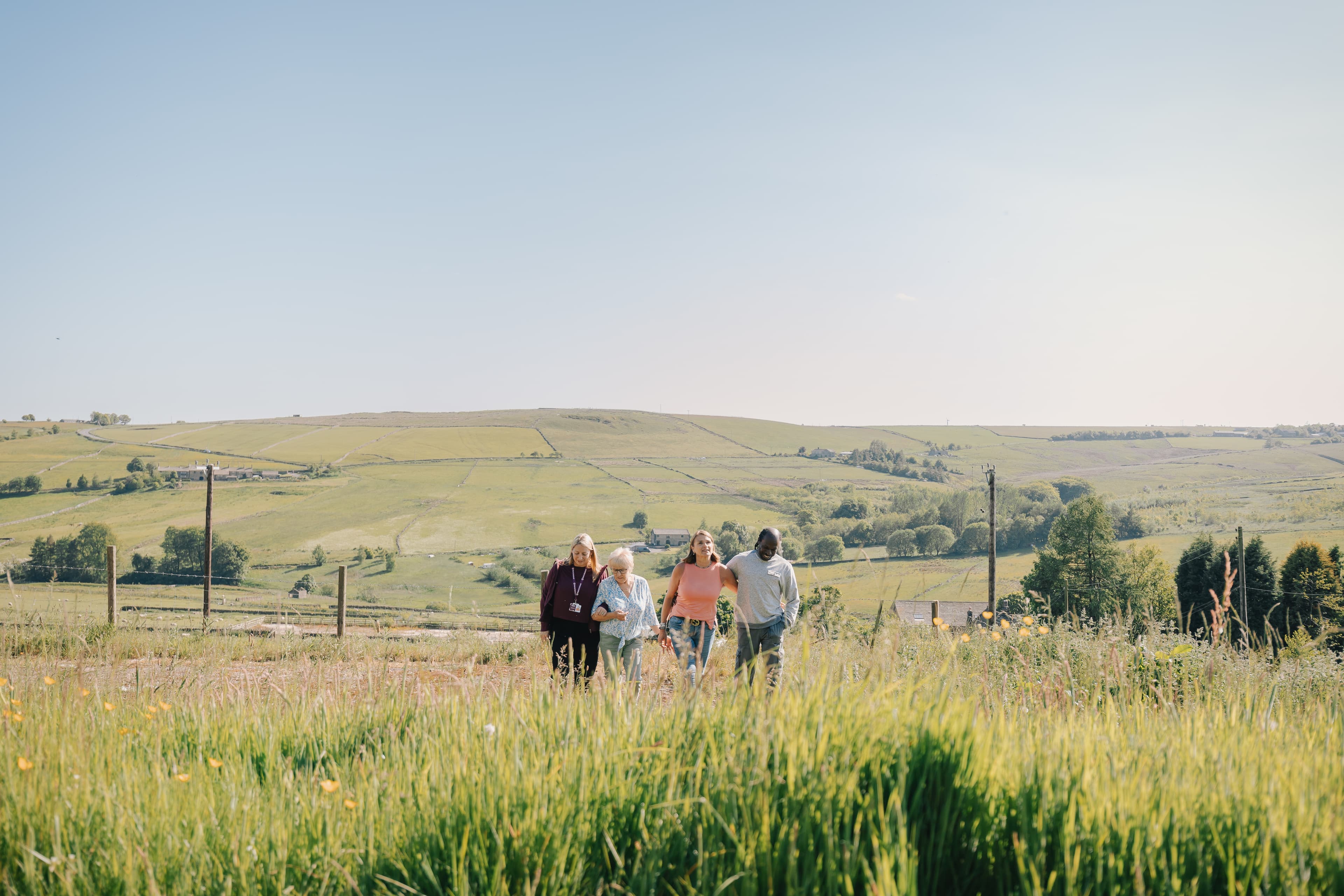 Four people walking together through a grassy field with hills and a clear sky in the background. - Home Instead