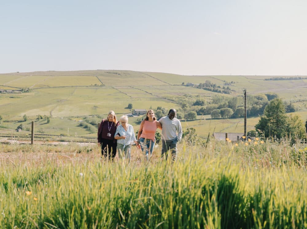 Four people walk together through tall grass in a countryside landscape with hills in the background. - Home Instead