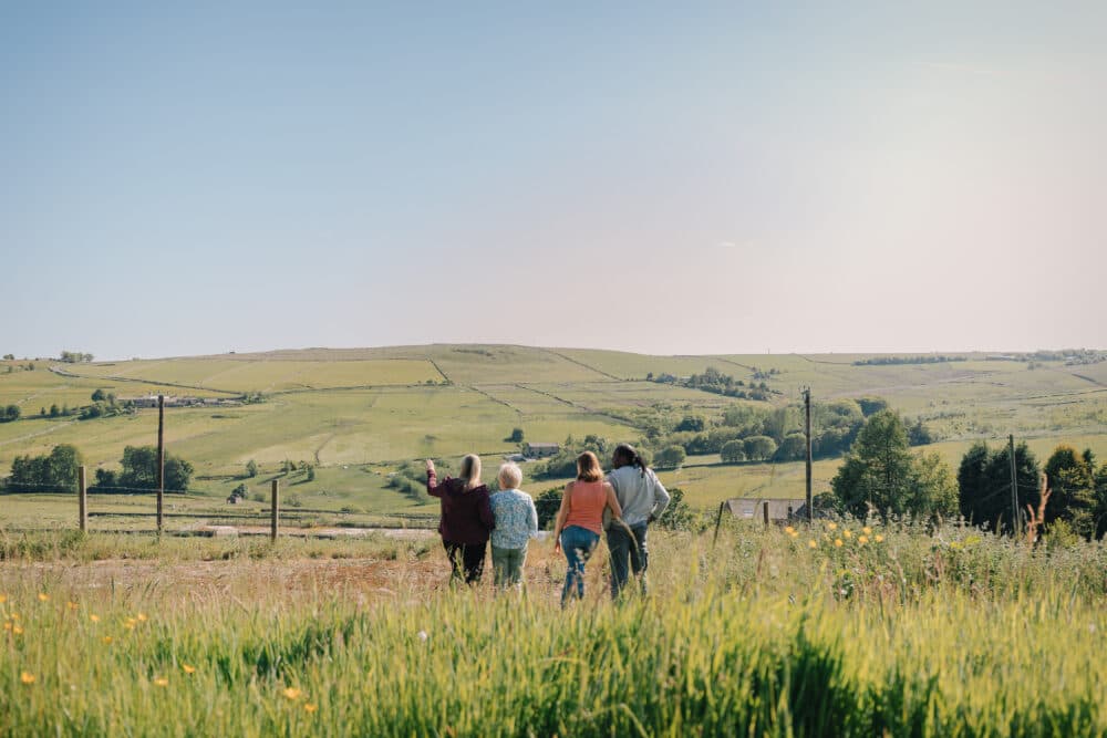 Four people stand in a grassy field, looking out at rolling green hills under a clear sky. - Home Instead