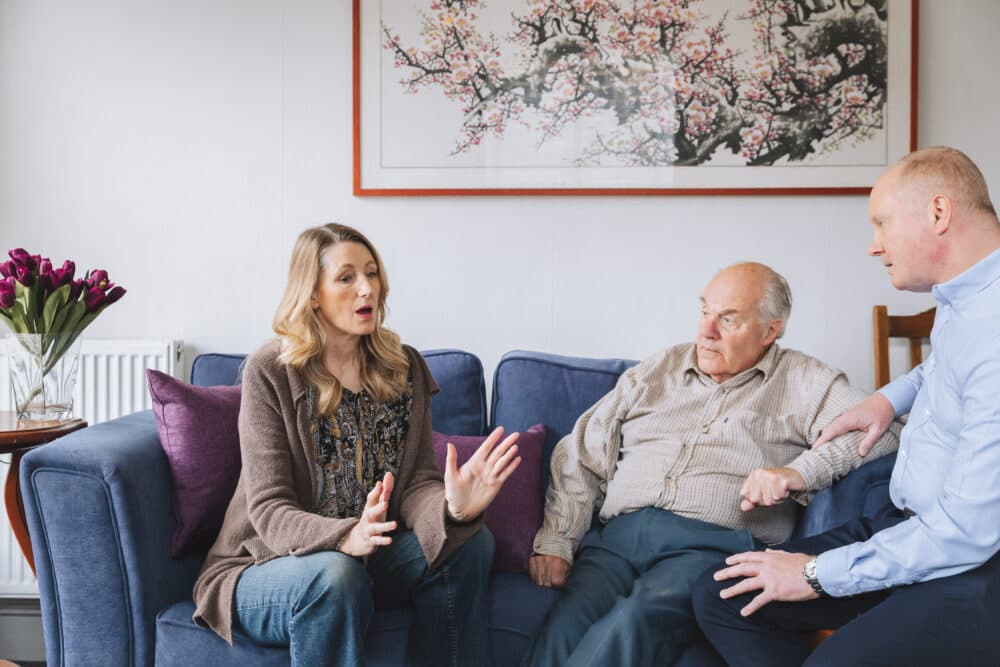 A woman talks to two men on a sofa in a living room with a painting and flowers in the background. - Home Instead
