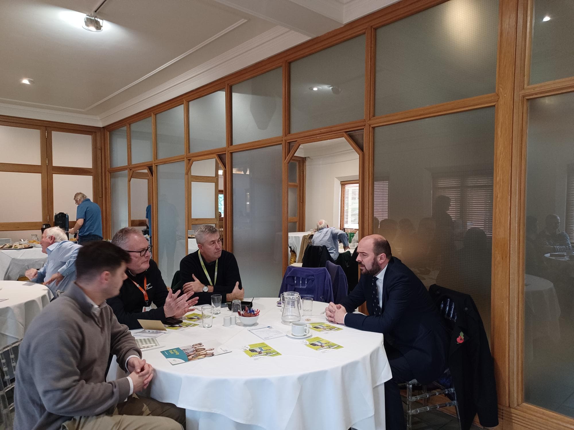 Four men sit around a table having a discussion in a conference or meeting room with glass dividers. - Home Instead