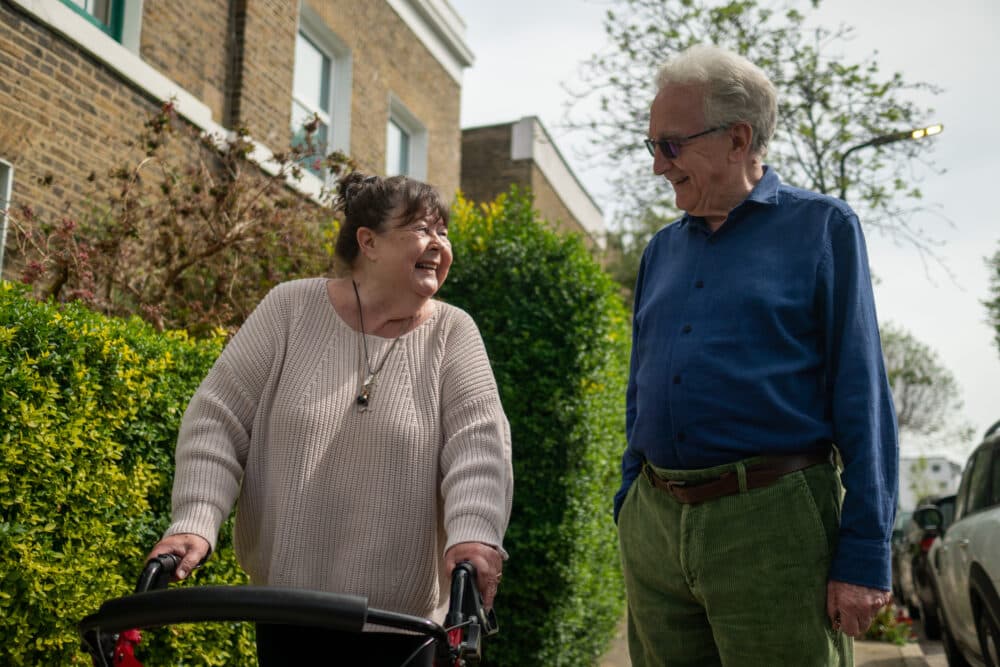 An older woman using a walker smiles at an older man as they talk outside on a sunny day. - Home Instead