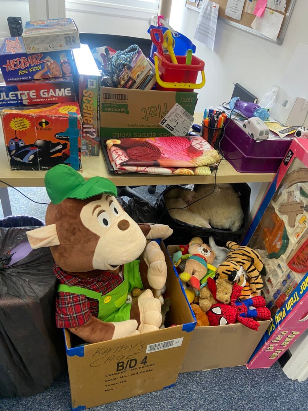 Boxes of assorted toys, games, and stuffed animals stacked near a desk in a cluttered room. - Home Instead