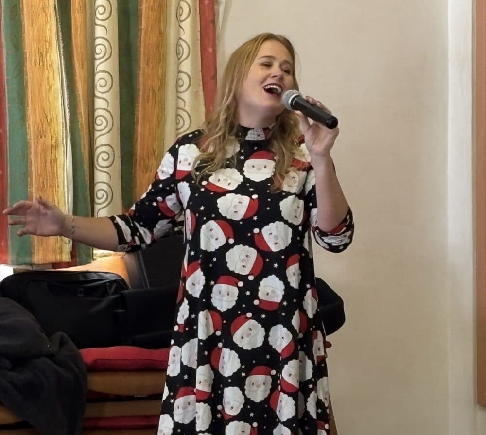 A woman in a Santa print dress sings into a microphone with one arm raised, standing indoors. - Home Instead