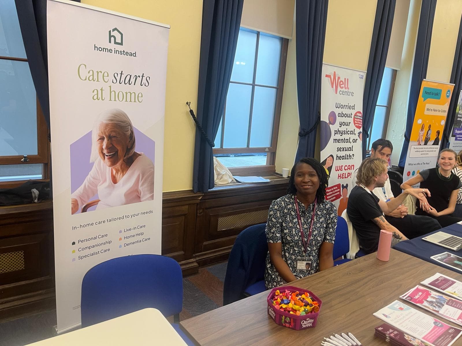 A woman sits at a table with leaflets and sweets, next to a "Care starts at home" banner at an event. - Home Instead