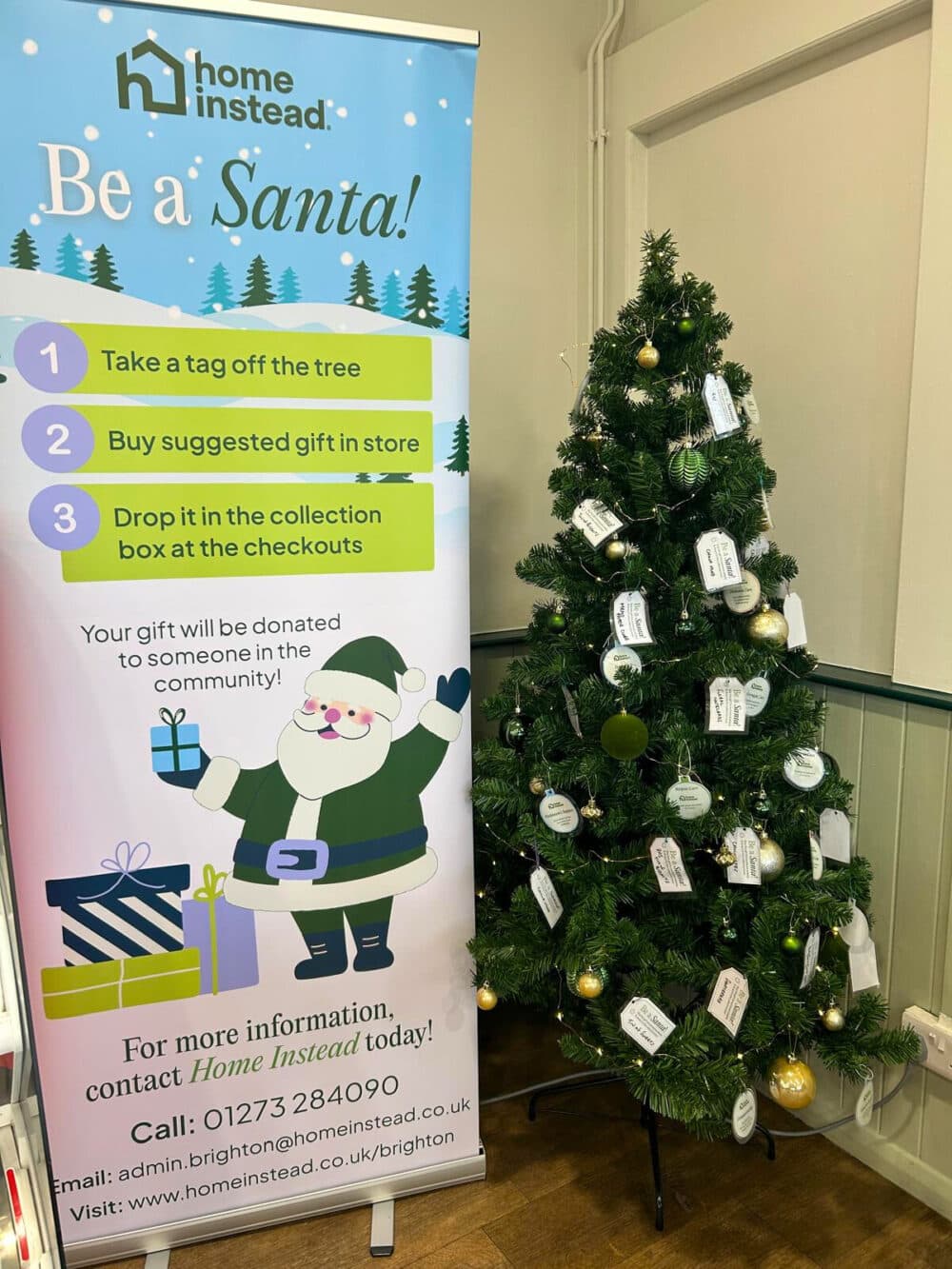 A Christmas display featuring a “Be a Santa!” banner from Home Instead next to a decorated Christmas tree with gift tags and ornaments.