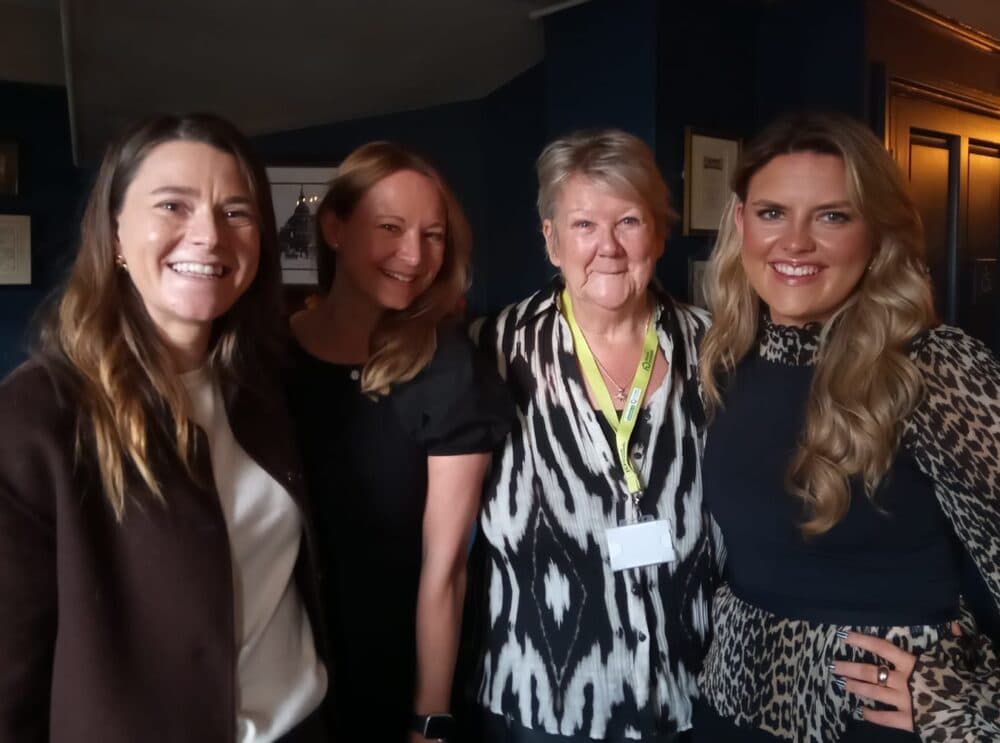 Four women standing together indoors, smiling at the camera, with dark walls in the background. - Home Instead