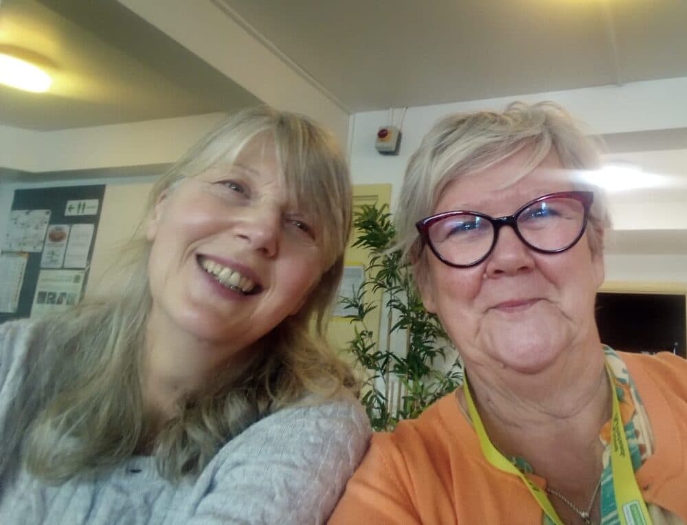 Two smiling older women indoors, one with glasses and a lanyard, posing for a selfie together. - Home Instead
