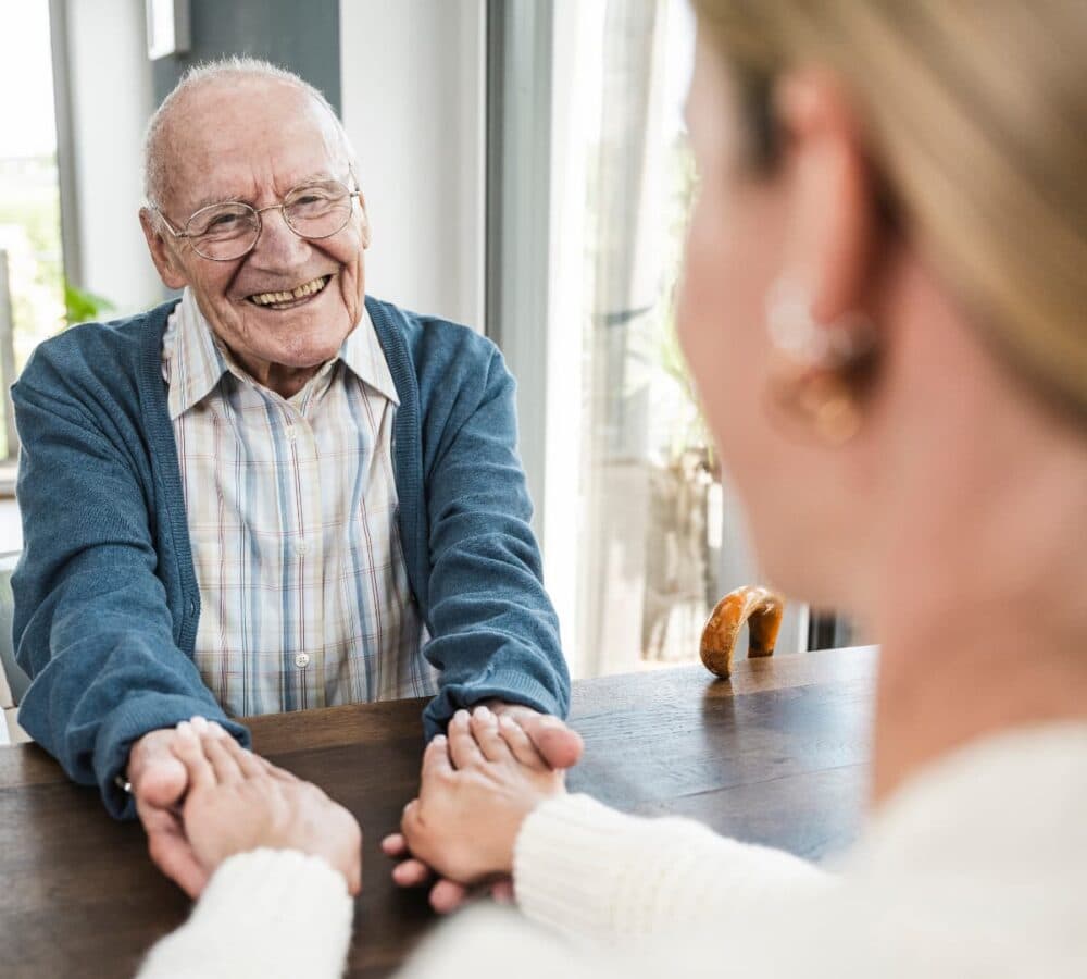 An elderly man smiles while holding hands with a woman across a table in a bright room. - Home Instead
