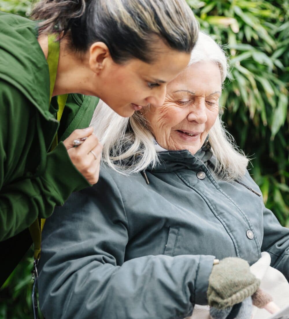 A young woman smiles and talks with an older woman who is sitting and wearing a green coat outdoors. - Home Instead