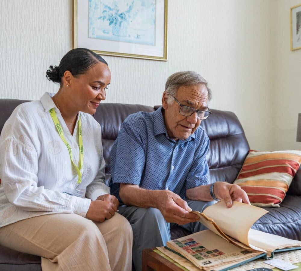 An older male adult with grey hair and wearing eyeglasses looking at an album while sitting on a couch with his younger female carer with black hair inside the house